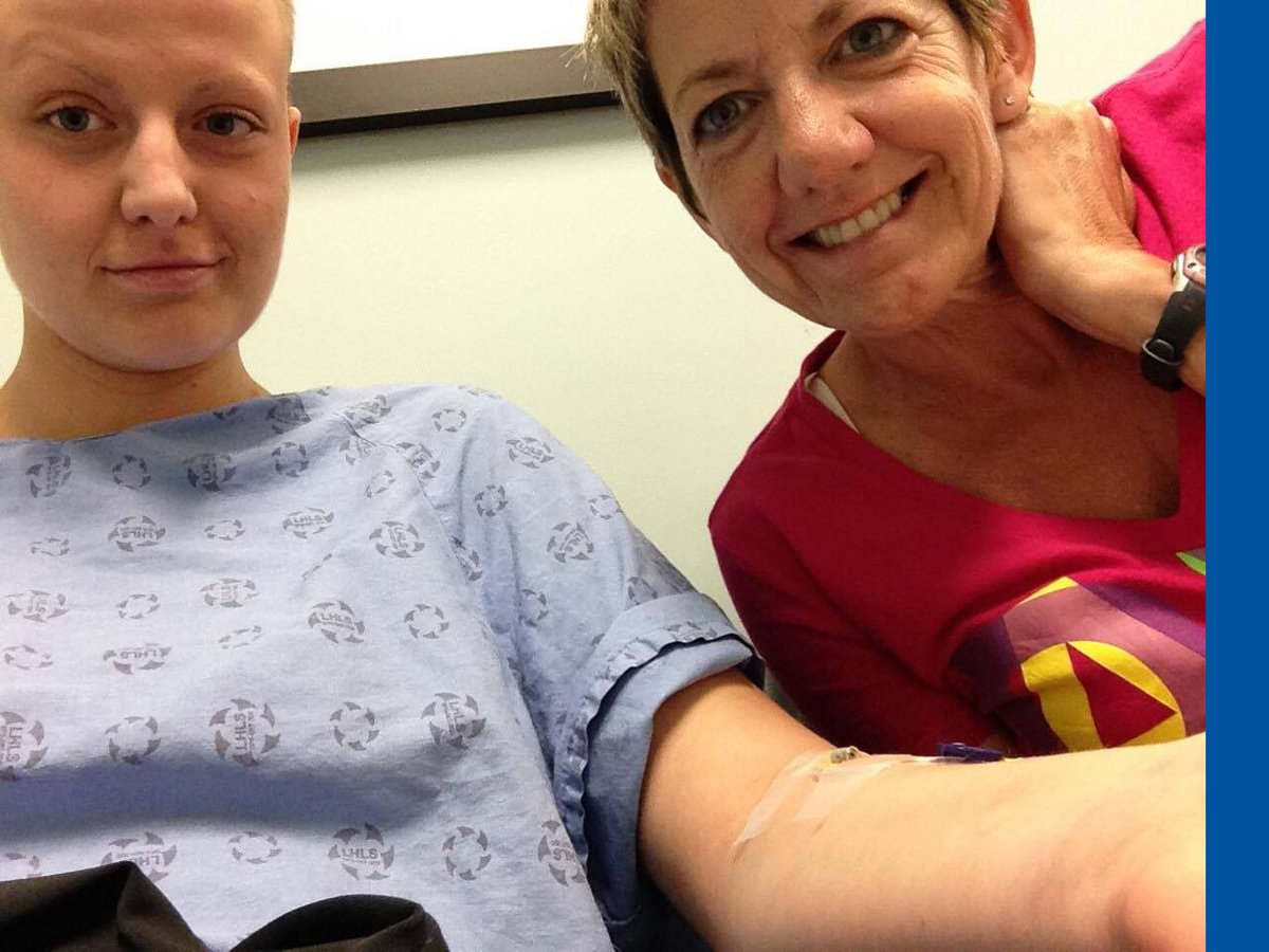 Emily, in a hospital gown with her head shaved, sits with her mom outside the CT suite before getting a scan.